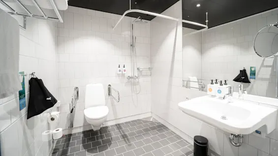 A bathroom with a white toilet, wall-mounted shower, and sink, all against tiled walls. A towel rack, toiletries, and a waste bin are visible. Bright overhead lighting. 