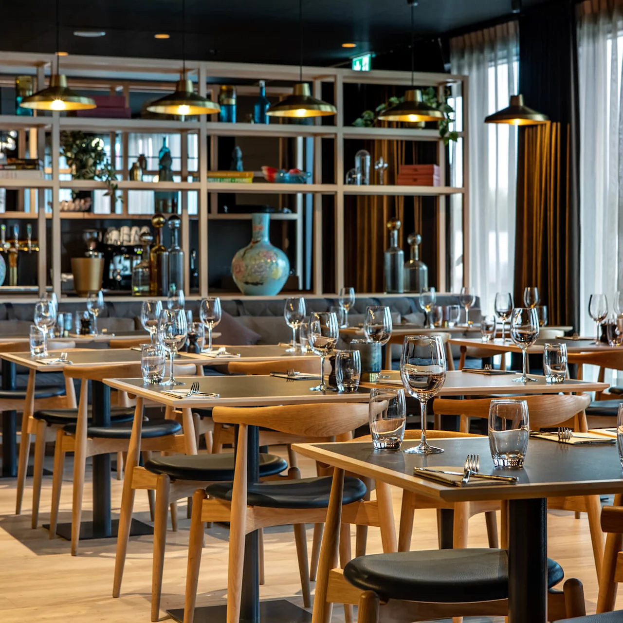 Luxuriously decorated restaurant interior, featuring neatly set wooden tables with glasses and cutlery. Elegant pendant lights hang above, and large windows provide natural light, enhancing the sophisticated ambiance.