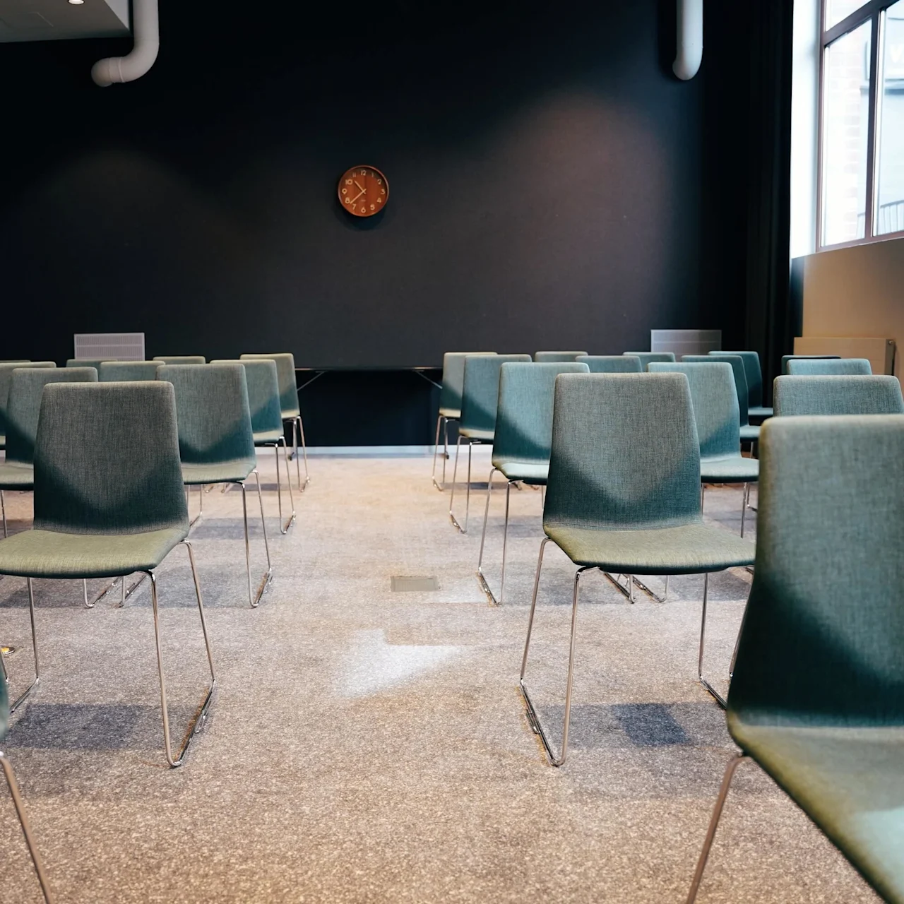 Rows of green chairs face a black wall with a brown clock, in a carpeted conference room with large windows and white curtains.