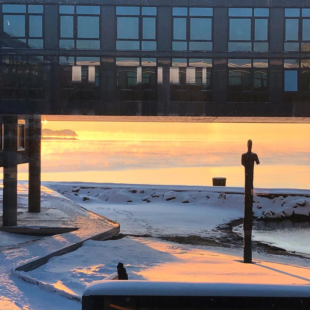En snedækket havnepromenade ved solnedgang med en statue, bygningsrefleksioner og en glødende lampe.