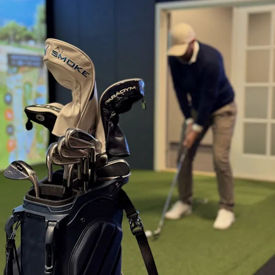 Golfbag med køller i forgrunnen og en golfer som trener på en innendørs golfsimulator i bakgrunnen.
