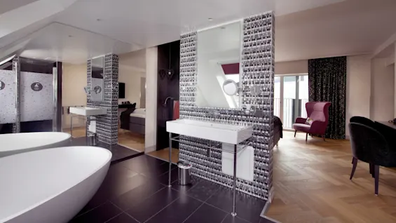 Freestanding bathtub sits on dark tile, near a mosaic-tiled sink area. Adjacent living space has wooden floors, a pink armchair, and a window with patterned curtains.