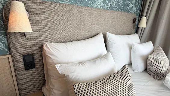 A cozy bedroom with a bed, white linens, and a nature-themed wallpaper. Wall lamps provide soft lighting.