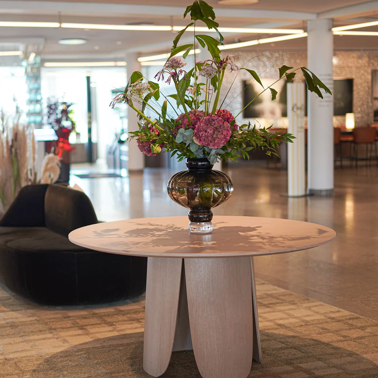 Et rundt bord har en stor blomsteroppsats med rosa og grønne blomster. Det står i en romslig, moderne lobby med elegante svarte stoler og dekorative planter i nærheten.