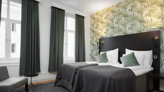A bedroom with two beds against a patterned wallpaper, flanked by a window with dark green curtains, and a gray upholstered chair.