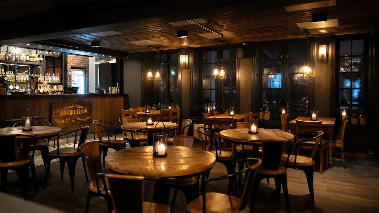 En koselig, dempet opplyst bar eller restaurant med trebord, stoler og tente lys. Bar synlig. Skilt: Trondhjems Biegte-Kro.