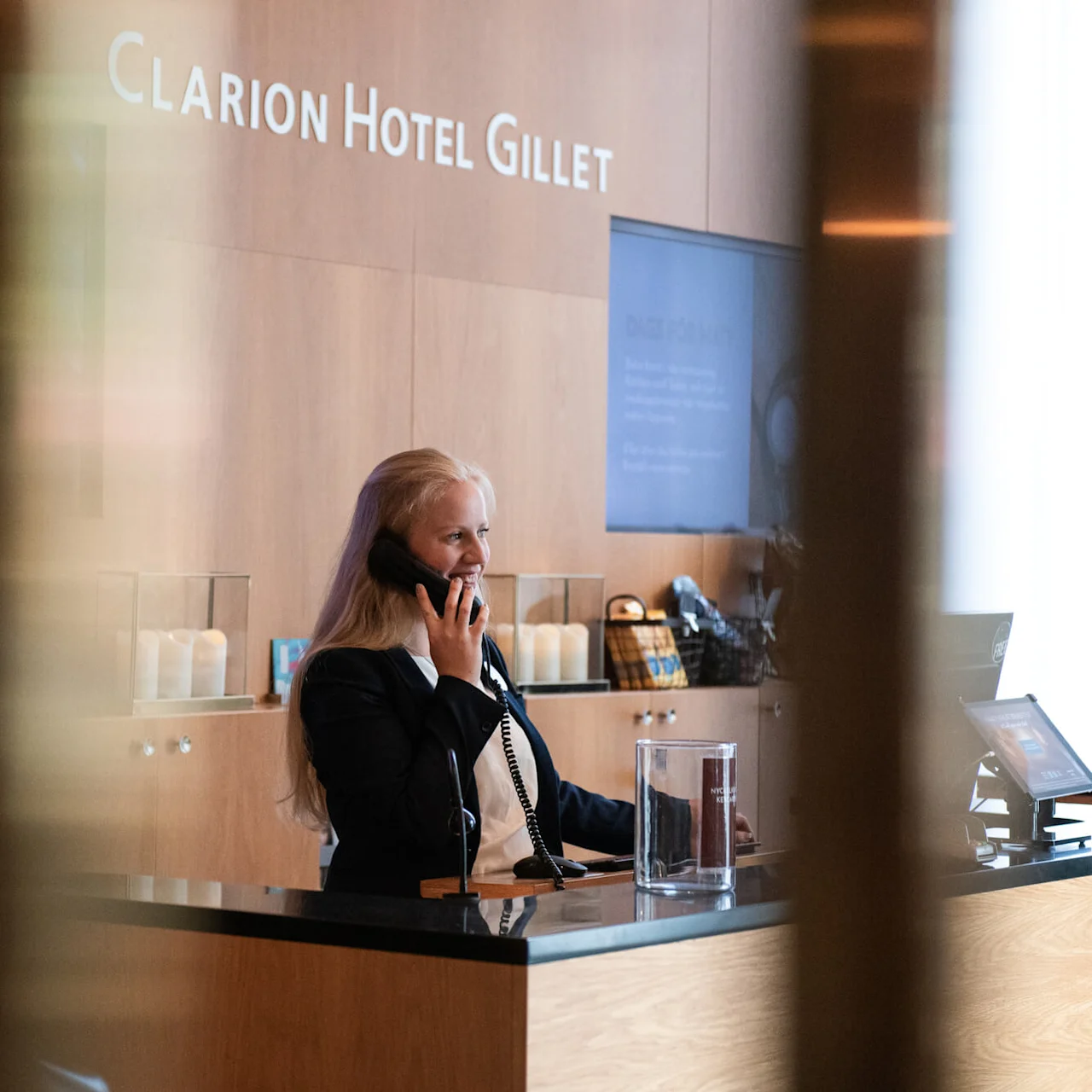 Woman on phone at hotel reception desk of Clarion Hotel Gillet; reception area features modern decor with wood paneling and candles, creating a welcoming ambiance.
