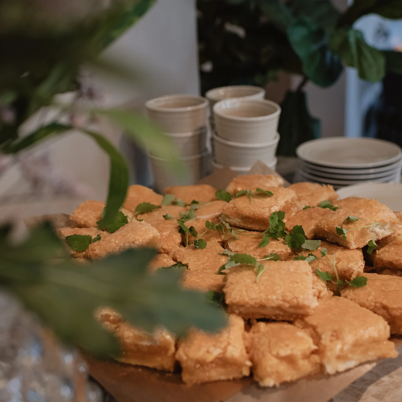 Golden brown pastries sprinkled with herbs are stacked on parchment paper; set on a table in a cozy room. Stacks of white cups and plates are in the background.
