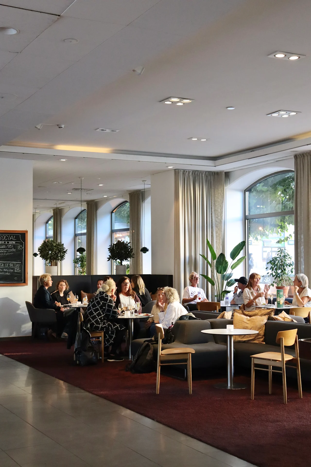 Café guests sit and socialize at tables, surrounded by plants and large windows in a well-lit, modern interior. A chalkboard menu on the wall reads: "SOCIAL FINNISH BEGINNER TIPS I DAY ON THE WAY."