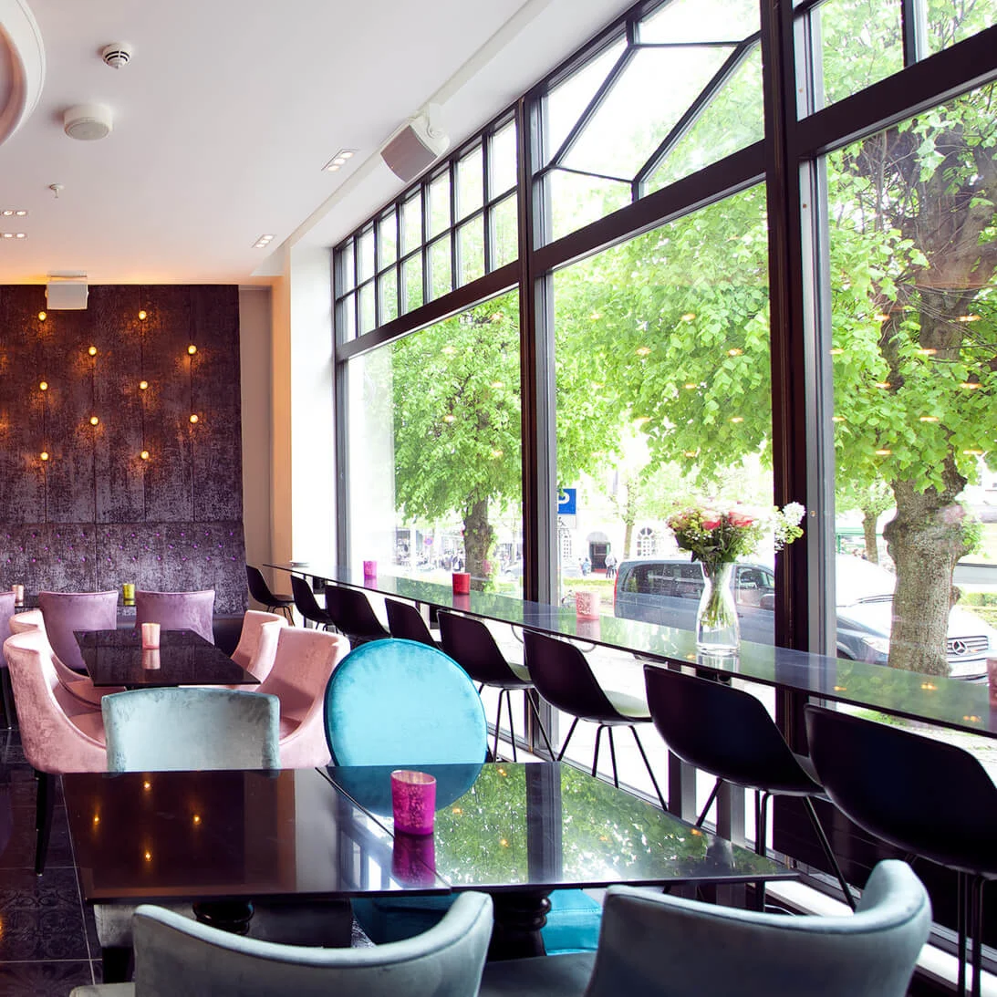 A large, purple chandelier hangs over a modern restaurant with colorful chairs and dark tables. Soft lighting decorates the far wall, and large windows reveal trees outside.