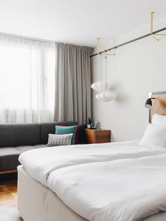 Moderne soveværelse med en pænt opredt seng, stilfulde lysarmaturer og et siddeområde med en grå sofa i et rent, lyst rum.