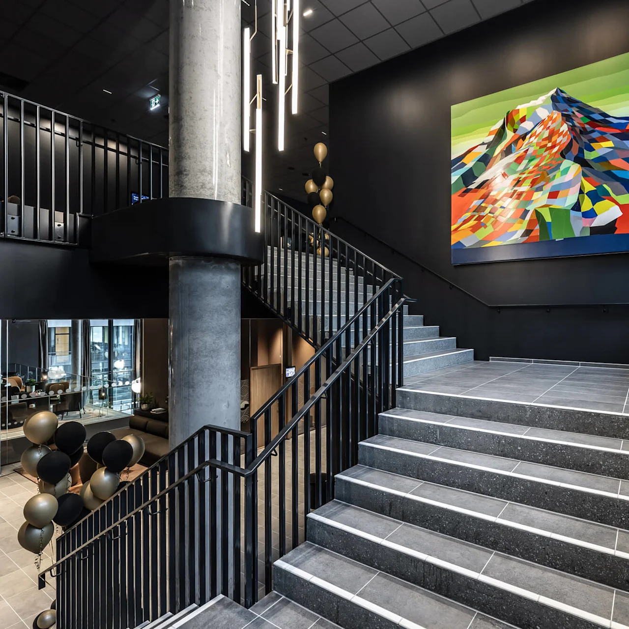 A large, colorful abstract painting hangs above a staircase in a modern interior. Balloons float nearby, and contemporary lighting accents the sleek, industrial design.