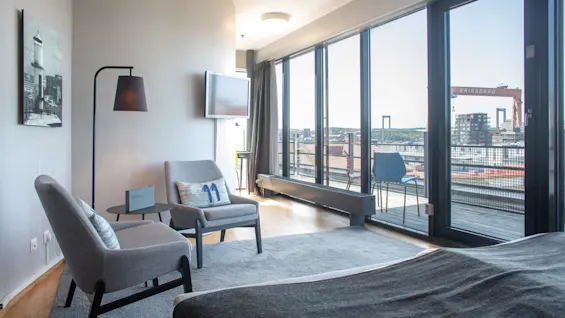 A room with modern gray chairs arranged by a small round table, facing large windows overlooking a city skyline with "ERIKSBERG" text on a crane outside. 