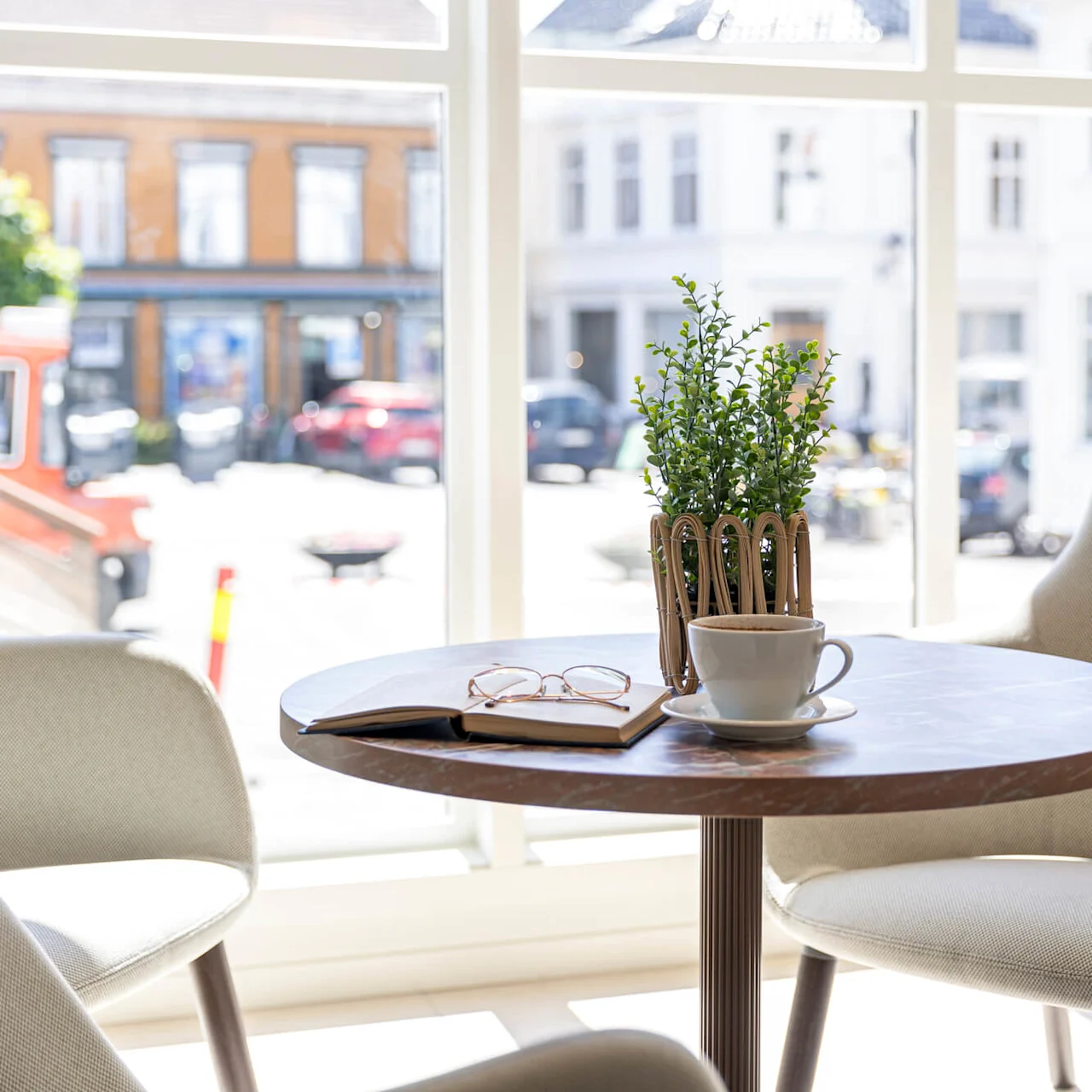 A coffee cup, glasses, and a plant on a table inside a cafe, with a street view outside.