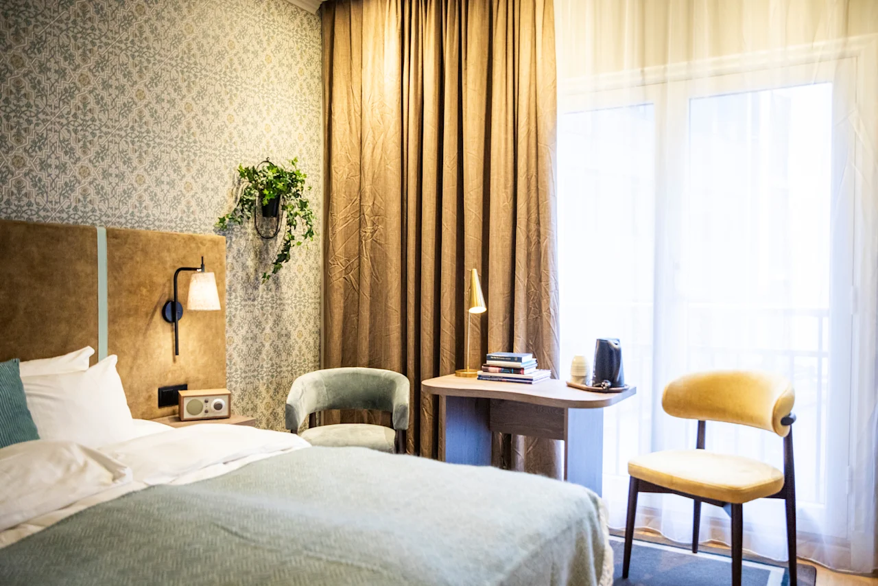 Säng med blå överkast bredvid ett nattduksbord med lampa, i rum med mönstrad tapet, stol, skrivbord och golvlånga gardiner nära ett fönster.