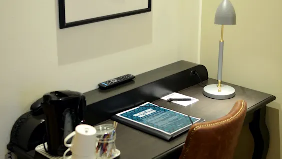 A desk with a lamp, phone, remote, notepad, pen, coffee cup, and glass holders in a room.