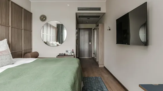 En pænt redt seng med grønt sengetæppe står i et moderne hotelværelse, med et vægmonteret TV og et rundt spejl overfor indgangsdøren.