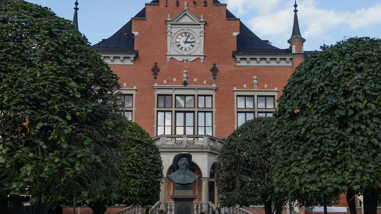 A grand brick building with a clock tower displaying 1898, a statue in front, flanked by trees and benches under a blue sky.