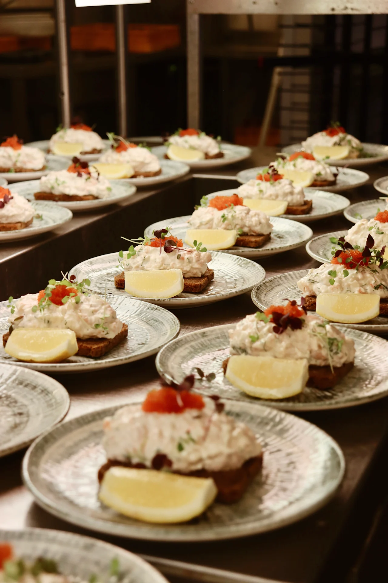 Open-faced sandwiches with creamy seafood salad and lemon wedges rest on ceramic plates, garnished with herbs, arranged in rows on a restaurant kitchen counter.