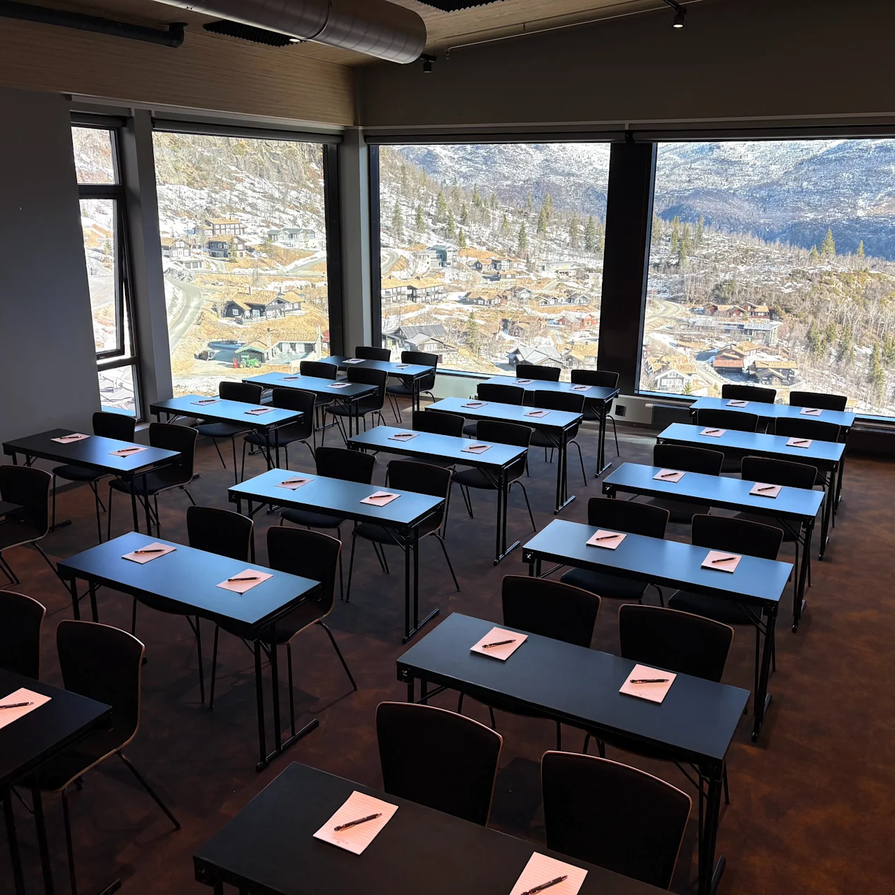 Classroom with neatly arranged black tables and chairs; notepads and pens are placed on each table. Large windows offer a view of a mountainous landscape with scattered buildings.
