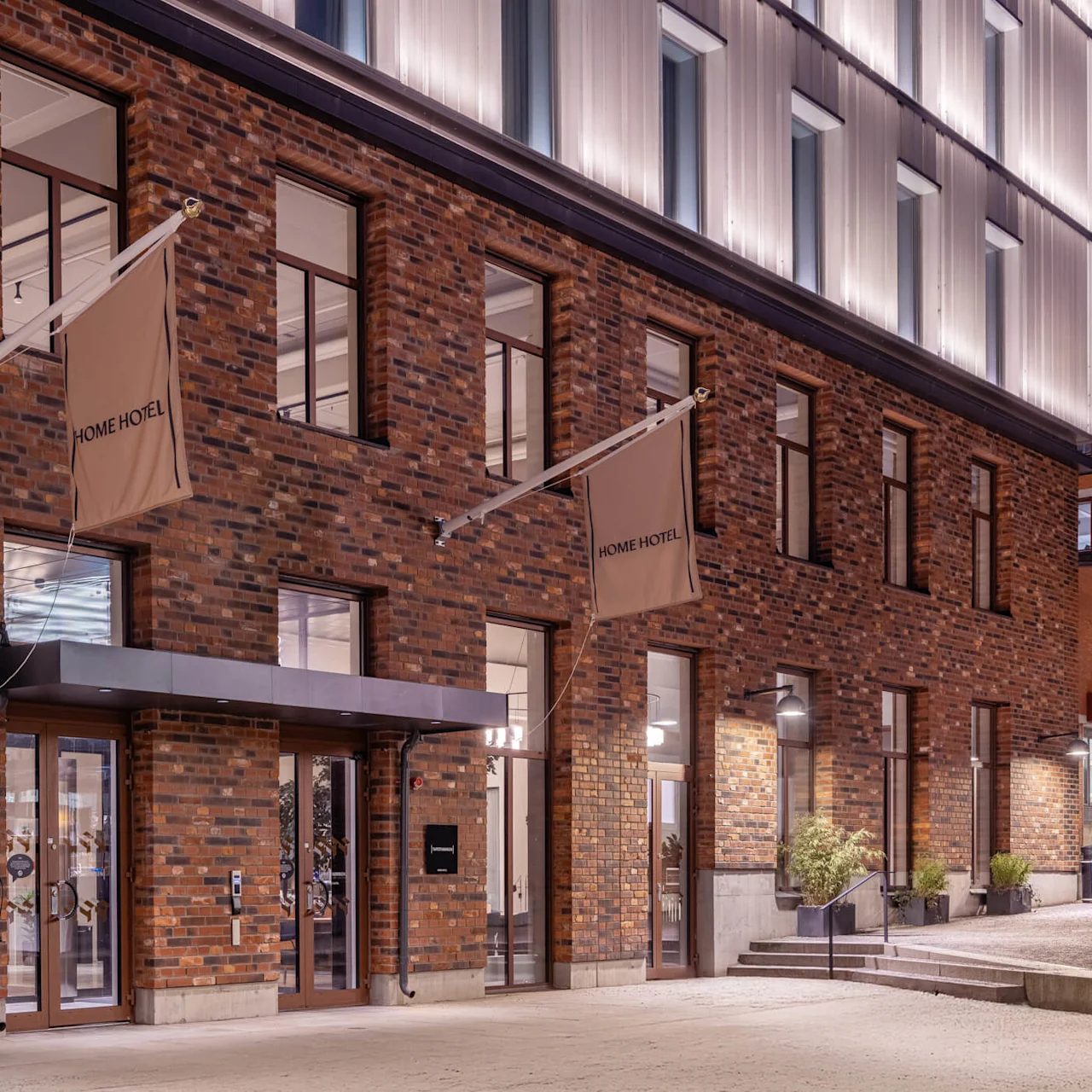 A brick building displays three beige flags with "HOME HOTEL" written on them, situated in an urban environment, featuring large windows and a lit entrance.