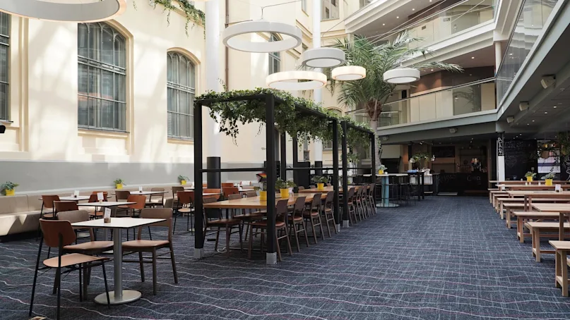 An indoor atrium cafe with tables, chairs, and a bar counter, sunlight streaming through arched windows, and plants adorning the space.