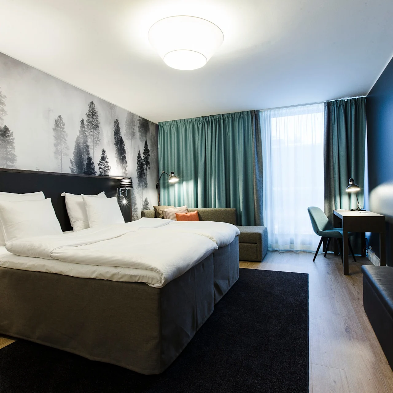 Bed with white linens and pillows, placed in a room with forest-themed wallpaper. Adjacent sofa and desk by a window with teal curtains; wall-mounted TV opposite the bed.