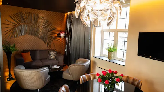 En lysekrone lyser opp en koselig, stilfull stue med en grå sofa, skinnstoler, et rundt svart bord med roser, dekorative planter og en veggmontert TV nær store vinduer.