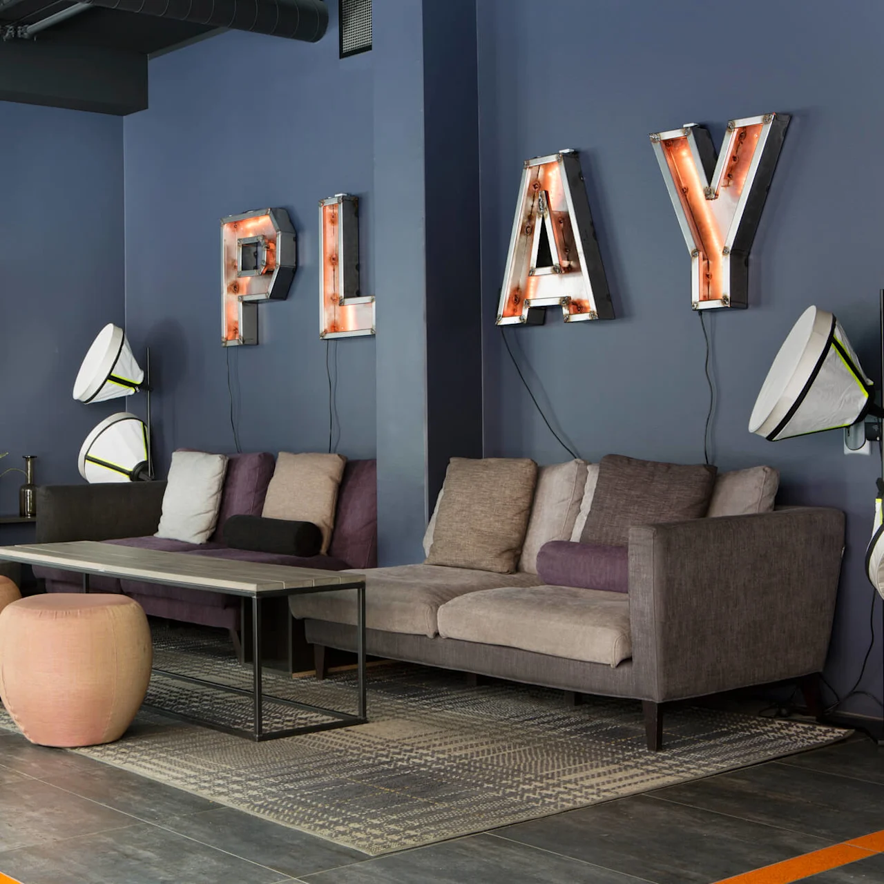 A cozy lounge area features a gray couch with cushions, a wall-mounted TV, and a decorative "PLAY" sign. Surrounded by chairs and plants, it's an inviting relaxation spot.
