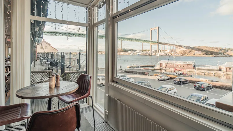 A round wooden table with two chairs overlooks a scenic waterfront view through large windows, with a bridge and buildings visible outside. Several cars are parked by the waterfront.