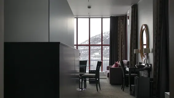 Et moderne og dæmpet belyst hotelværelse med møbler, med udsigt til snebeklædte bakker og vand gennem store vinduer.