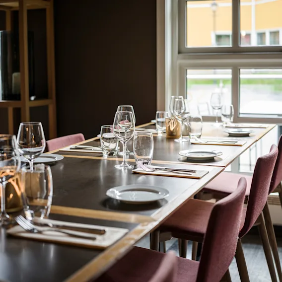 Spisebord dekket med vinglass, tallerkener og bestikk på servietter; dekorert med lys. Omgitt av røde stoler ved store vinduer som slipper inn naturlig lys i en koselig restaurant.