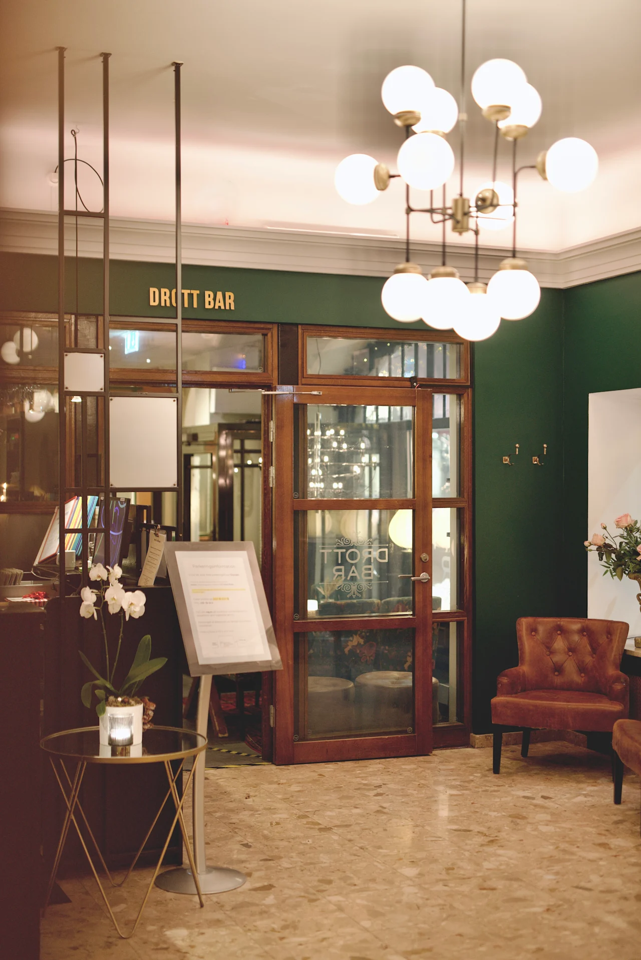 En ljuskrona hänger i en hotellobby med marmorgolv, bruna stolar och orkidéer i krukor. En skylt lyder "DROTT BAR" ovanför en dörr av glas.