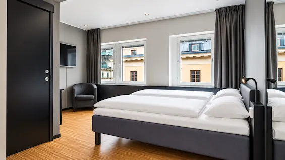 Bright hotel room featuring a made double bed, wooden flooring, and windows overlooking the city.