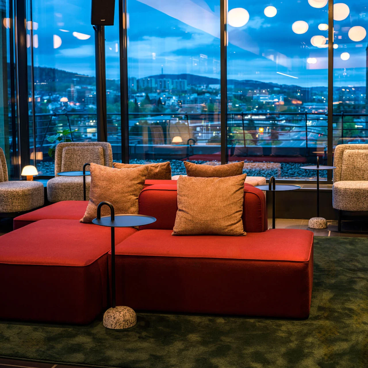 Modern lounge features red modular sofas with cushions, surrounded by gray chairs and small tables, set against large windows with an evening cityscape view, under warm ceiling lights.