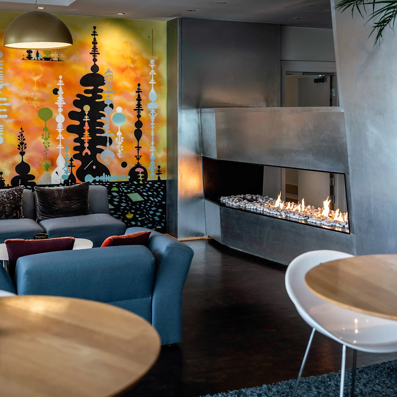 En färgglad väggmålning dekorerar en modern lounge med blå soffor och en snygg öppen spis. Träbord och mjuk belysning skapar en mysig atmosfär.