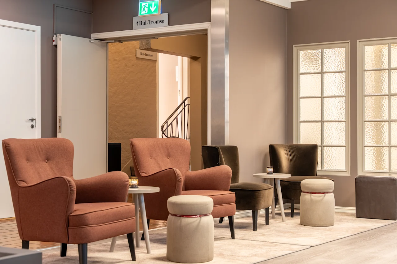 A comfortable hotel lounge with armchairs and small tables. An exit sign points to Bul-Tromso, leading to a staircase.
