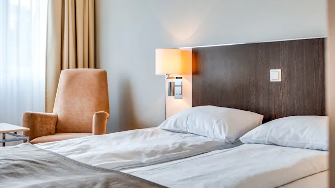 A neatly made bed with a tan armchair beside it, illuminated by a lamp, in a calm hotel room.