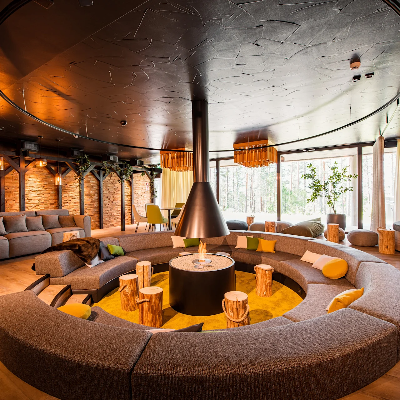 A circular gray sofa surrounds a hanging fireplace in a modern lounge with wooden accents and large windows displaying greenery outside.