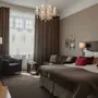 Ett mysigt hotellrum har en stor säng med vinröda kuddar, en ljuskrona ovanför och en sittgrupp med stolar vid ett gardinbeklätt fönster; varm belysning förstärker den inbjudande atmosfären.