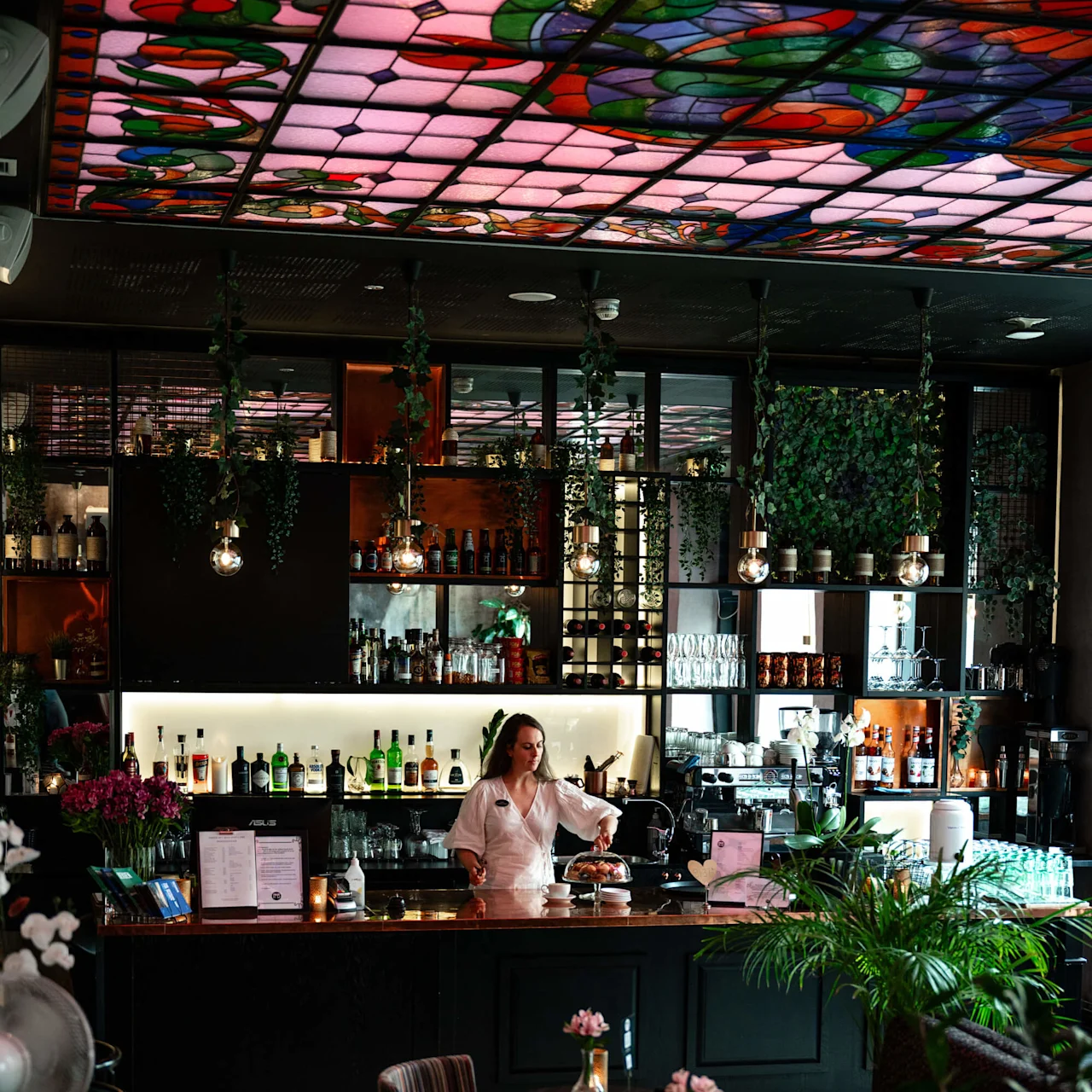 Bartender tilbereder drikkevarer bak en koselig bar med hengende planter og farget glassloft. Stemningslys reflekterer fra de mørke møblene. Små runde bord er pyntet med rosa blomster.