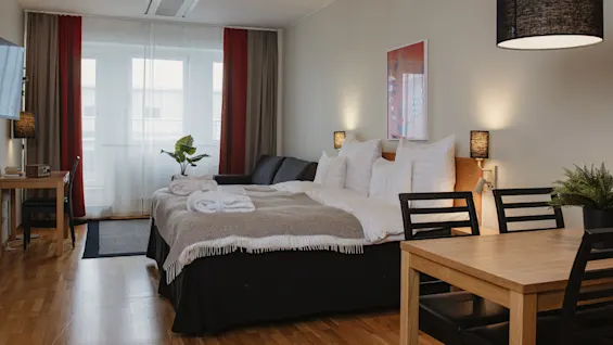 A neatly made bed with white pillows and a beige throw, positioned beside a small table and chairs, in a well-lit room with wooden flooring and large windows.