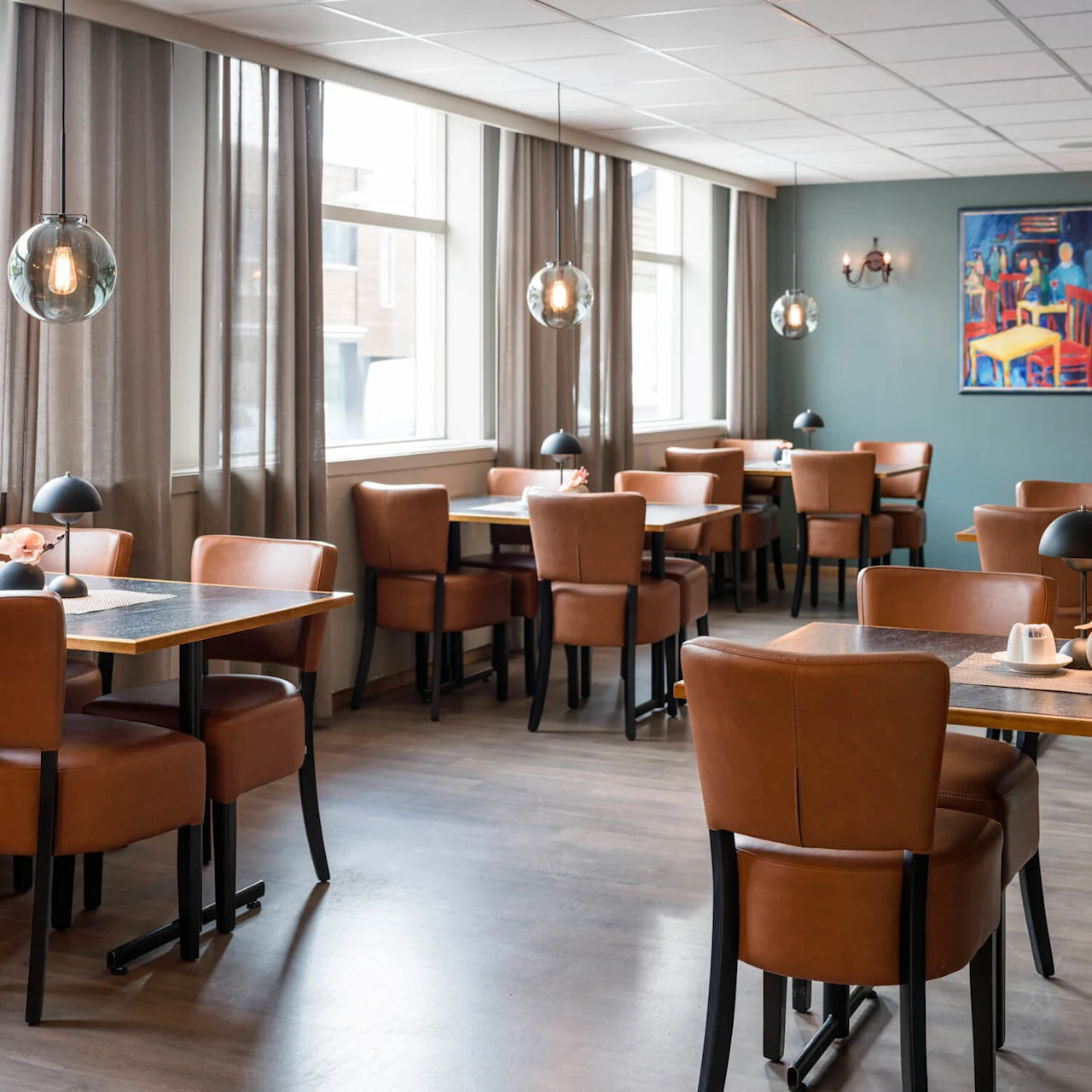 Tables and chairs sit in an empty, well-lit restaurant. Leather chairs, pendant lights, and a colorful painting on the wall define the elegant setting.