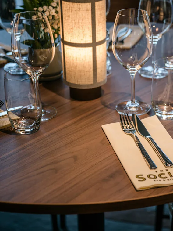Et rundt trebord dekket for middag med vinglass, bestikk på servietter merket "THE SOCIAL BAR & BISTRO," rundt en mykt opplyst lampe. Elegant, koselig restaurantstemning.