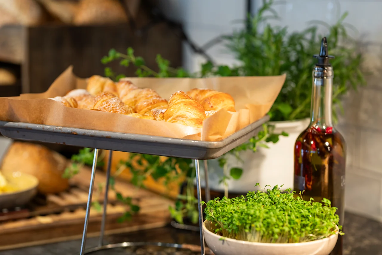 Croissanter vilar på en bakplåt med bakplåtspapper. I närheten står en flaska olivolja och krukväxtörter på en köksbänk och skapar en fräsch matlagningsatmosfär.