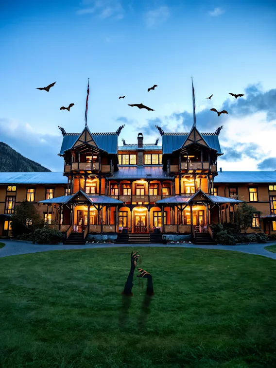 En storslået, oplyst herregård står med flagermus der flyver over i skumringen, omgivet af frodige haver og bjerge. Hænder dukker op fra græsset i forgrunden.