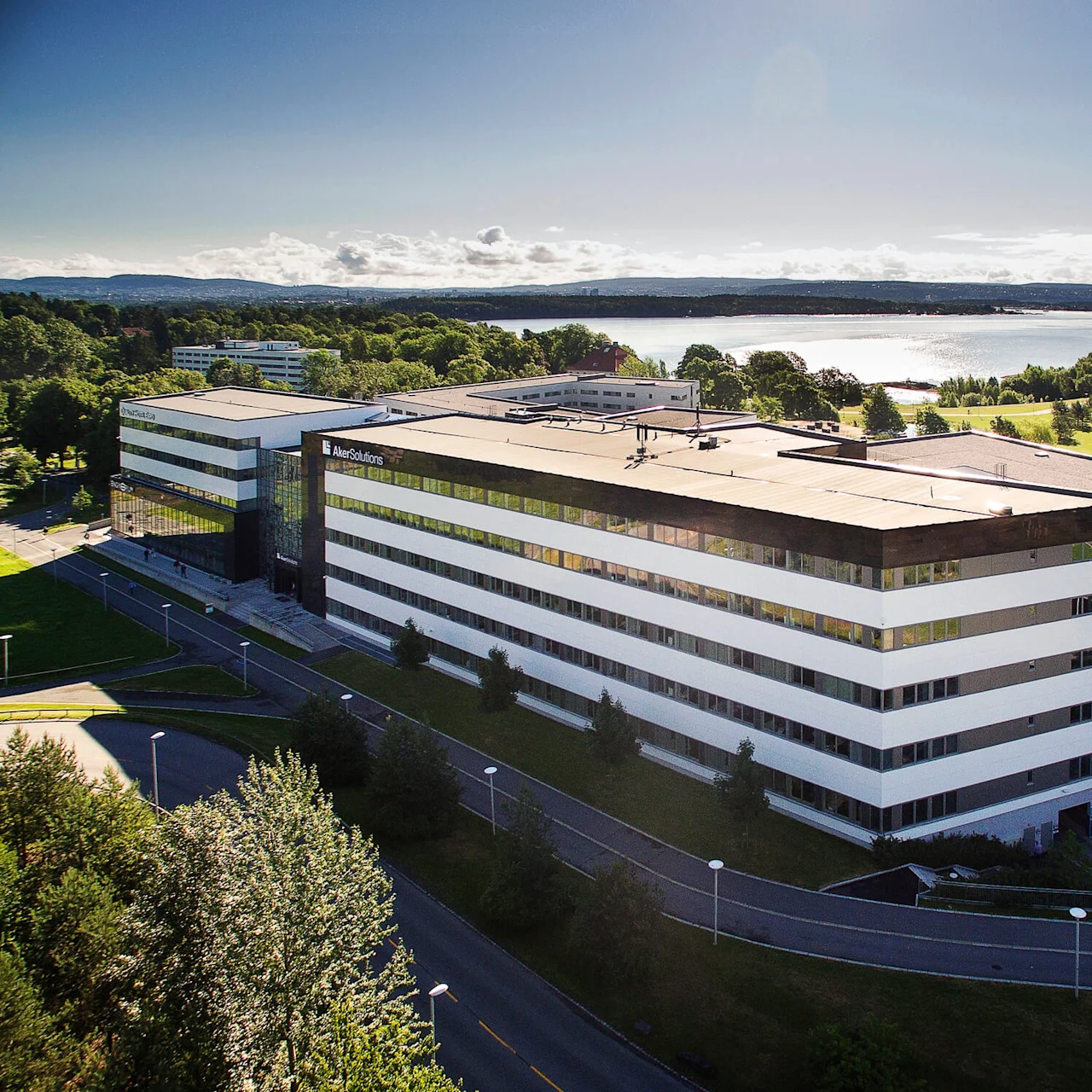 A large, modern building with "Aker Solutions" on the facade stands amid green lawns and trees, near a body of water under a clear blue sky.