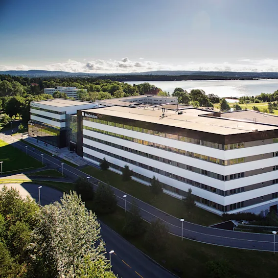 En stor, moderne bygning med "Aker Solutions" på fasaden står blant gressplener og trær, nær et vannlegeme under en klar blå himmel.