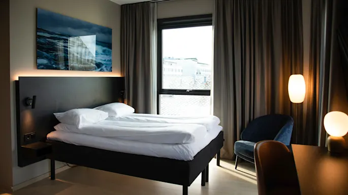 A neatly made bed with white linens is positioned in a modern bedroom, featuring large windows, dark curtains, blue armchair, and soft lighting from lamps. A seascape painting hangs above.