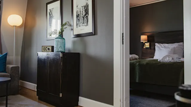 A dark wooden cabinet with a vase and clock sits next to a lit floor lamp; through an open doorway, a bedroom with a neatly made bed and wall lamp is visible.